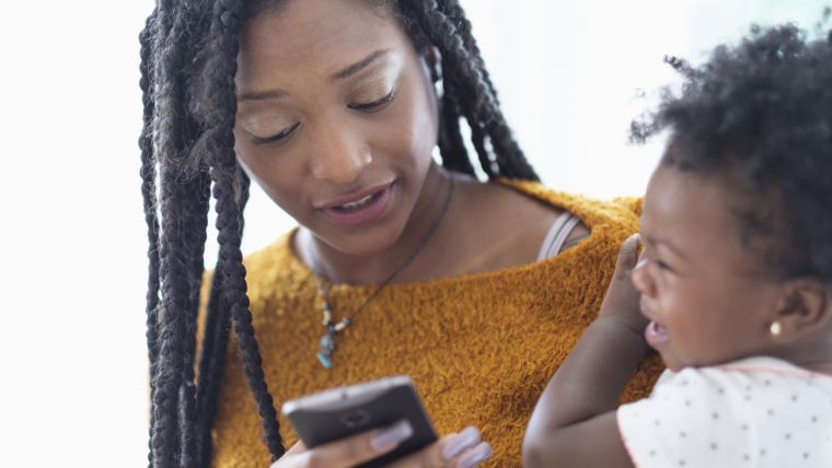 Black woman holding baby daughter using cell phone